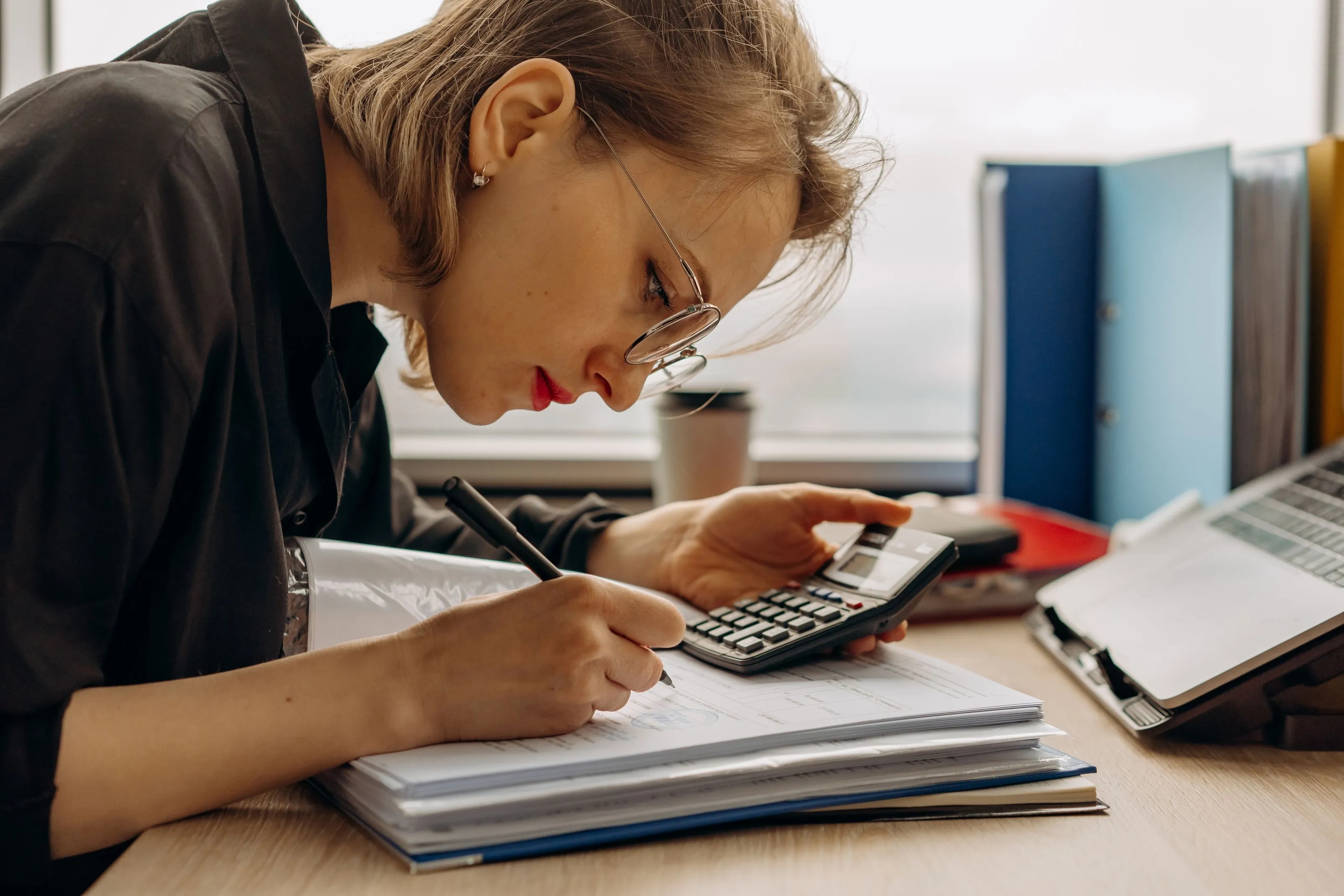 A woman in glasses writes figures in a binder while working a calculator at a desk by the window.