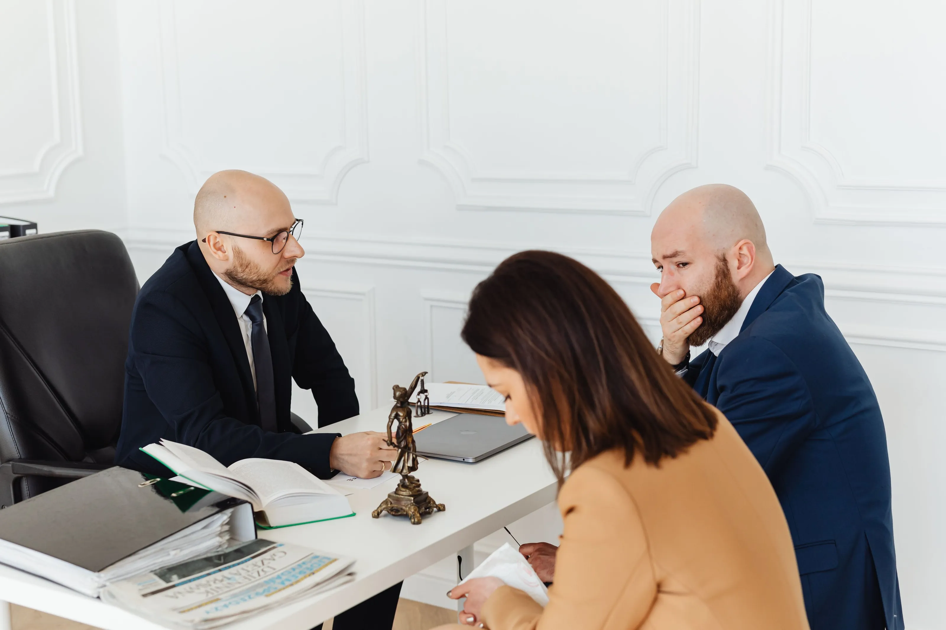 An attorney across a desk from two clients. One client reads a document while the other listens, hand over mouth.